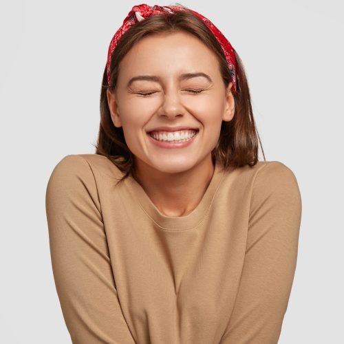 Portrait of happy European woman has broad smile, closes eyes, feels excitement, being in high spirit, recieves proposal from boyfriend, laughs at something funny, models against white background
